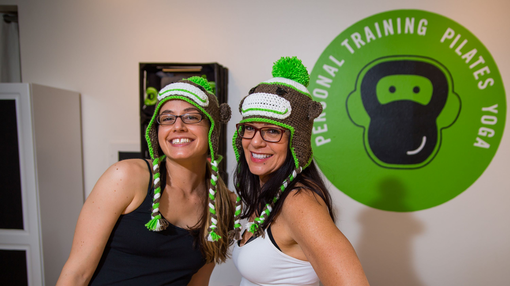 Kelly and her mom standing side by side in the Fit Monkeys studio, smiling and wearing crocheted monkey hats.